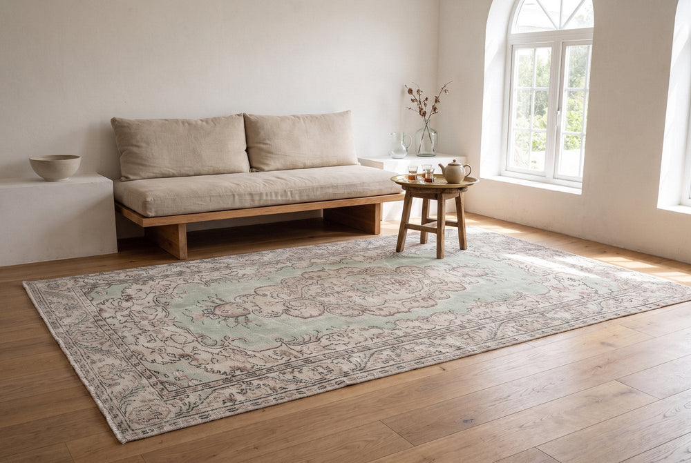 A sage green vintage Manisa rug with a pale medallion in a sunlit whitewashed room, with a low linen daybed and a brass tray of Turkish tea glasses on a wooden stool