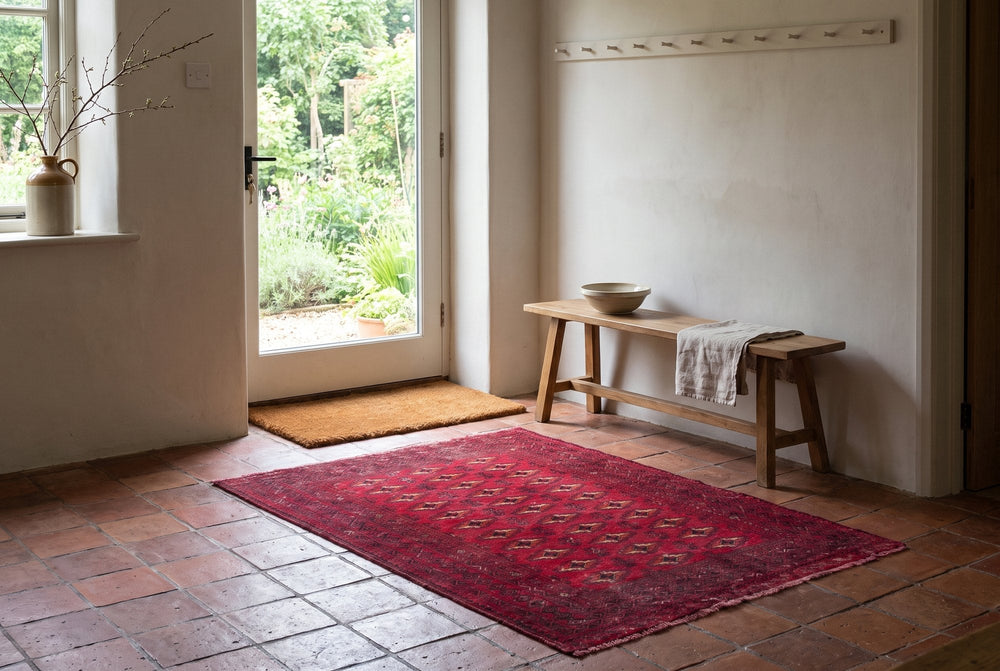 A small red Baluch rug with rows of gul motifs on a terracotta tile floor inside a glazed garden door, beside a wooden bench