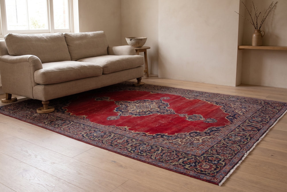 A deep red vintage Arak rug with a navy medallion and border under a linen sofa whose legs stand on round wooden furniture coasters, on a pale oak floor