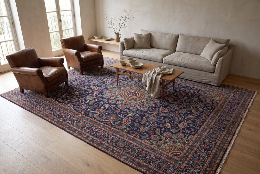 A navy Kashmar rug with dense vines, a round medallion and a rust and navy border under a linen sofa, two leather armchairs and an oak coffee table, seen from a high corner