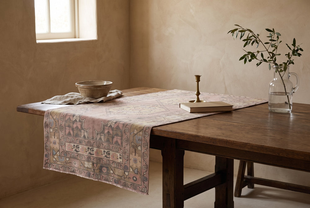 A dusty rose vintage Anatolian runner draped over a dark oak table like a Renaissance table carpet, with a brass candlestick, a plain book and a stoneware bowl