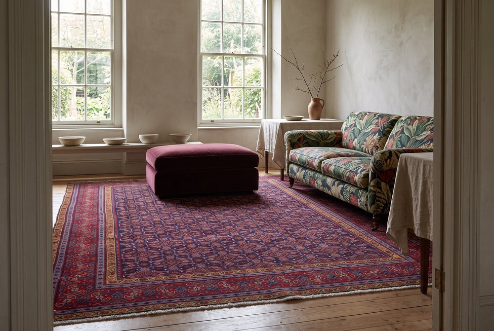 A navy vintage Mahal rug with a small all-over repeat and a red border beside a sofa in a bold botanical print and a plum velvet ottoman