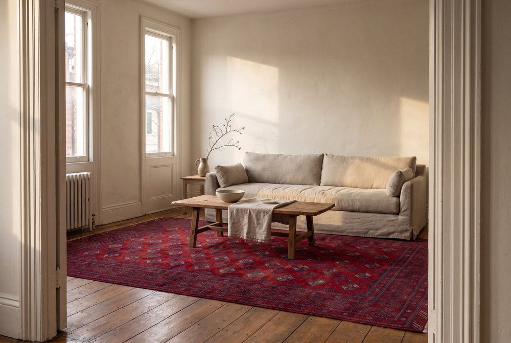 A large deep red vintage Torkaman rug with rows of small guls spread across a wooden floor in front of a linen sofa and a rough oak coffee table, seen through a doorway