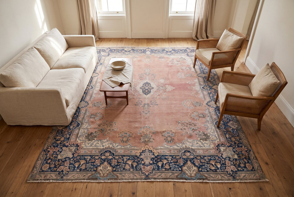 A muted pink Kerman rug with a navy border holding a linen sofa, two cane armchairs and a low walnut table, seen from high in the room