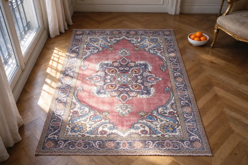 Looking down at a dusty rose Turkish rug with a navy medallion and cream corners on oak herringbone parquet, morning sun from tall windows
