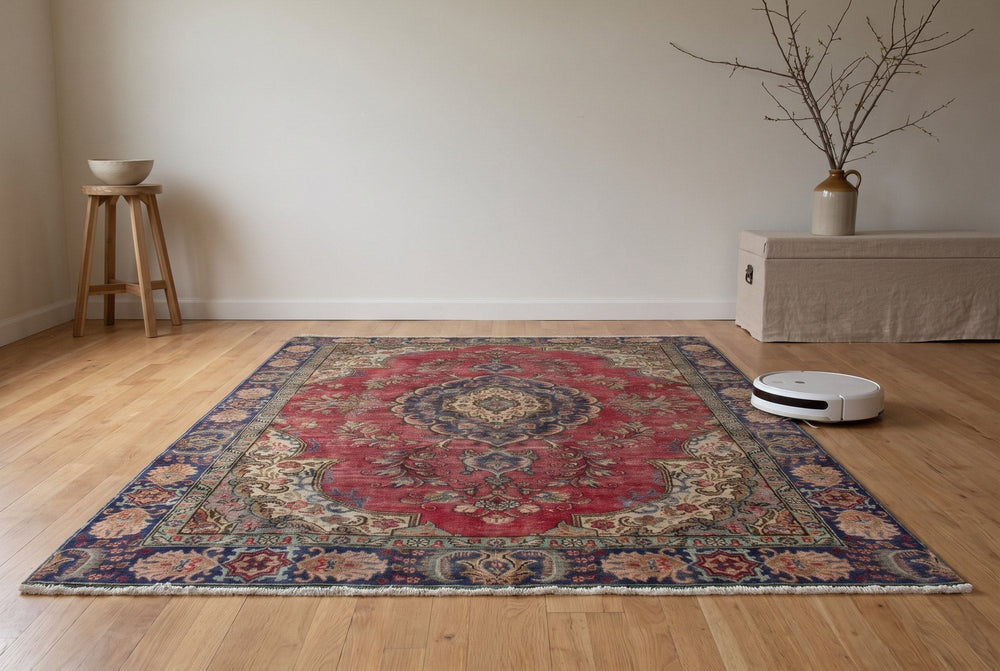 A deep red vintage Tabriz rug with a navy medallion and floral border on an oak floor, with a plain white robot vacuum parked just off its edge