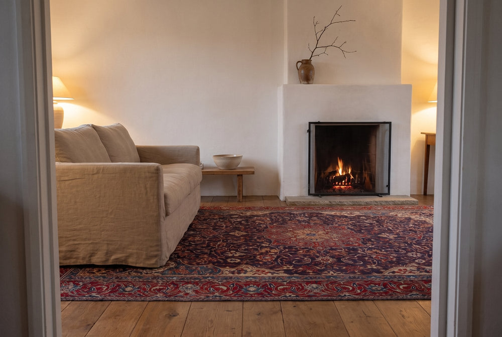 A navy vintage Mahal rug with a red medallion and red floral border under a linen sofa, a stone hearth and a screened fire beyond it, in warm evening lamplight