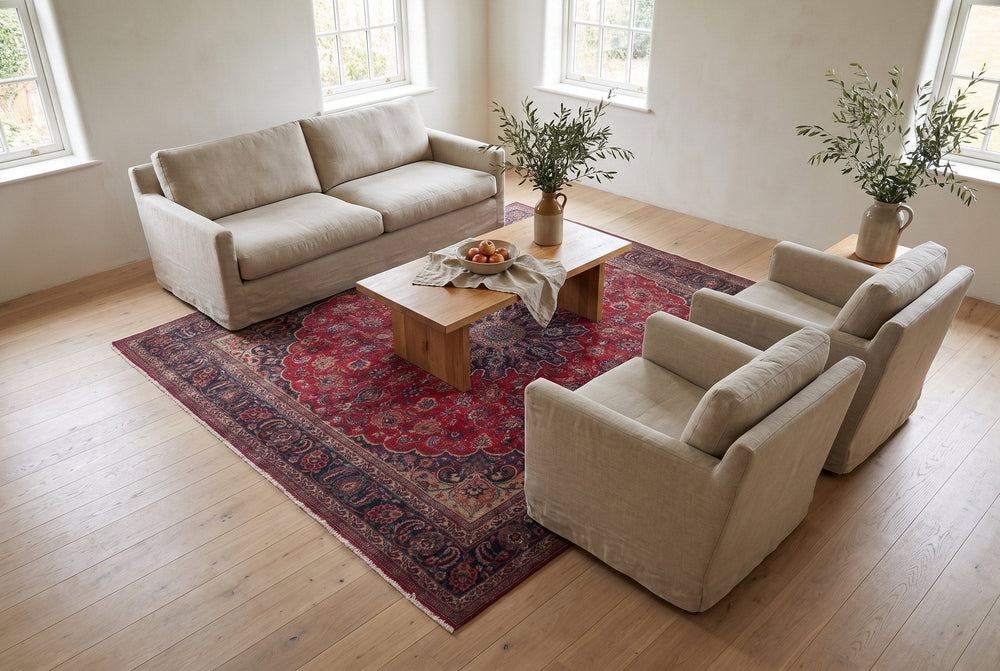 A square deep red vintage Sabzevar rug with a round floral medallion and navy border holding a linen sofa, two armchairs and an oak coffee table in a bright room