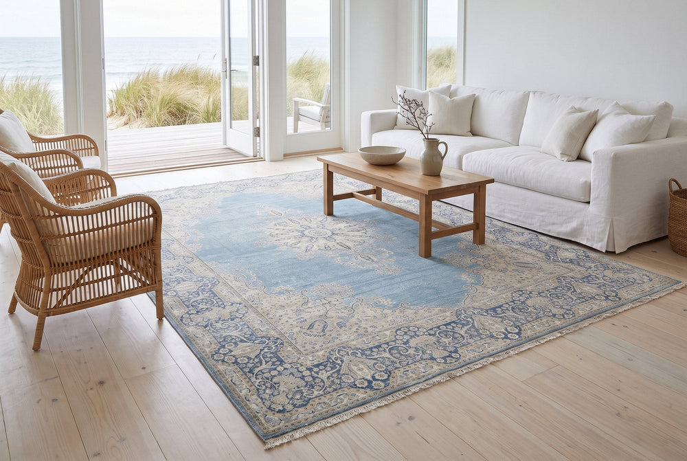 A powder blue vintage Kerman rug with a cream medallion and navy floral border in an airy coastal living room with a white linen sofa, rattan chairs and glass doors open to the dunes