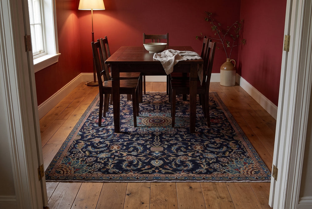 A navy vintage Kashmar rug with a turquoise and ochre medallion and scrolling vines under a dark wood dining table in a room with deep red walls, seen through a doorway