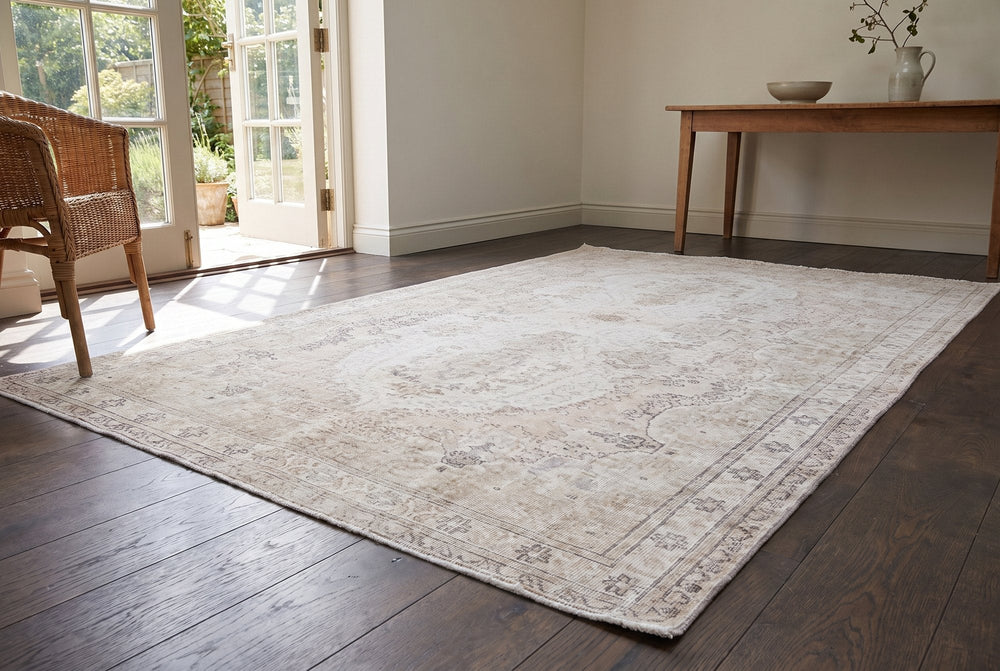 A pale cream vintage Isparta rug with a faint medallion airing on dark smoked oak boards in a garden room with French doors open and a wicker chair