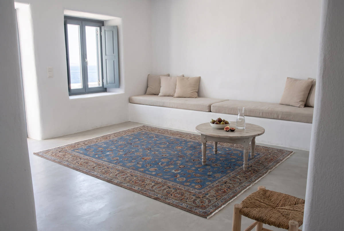A vintage Persian rug with a sky blue field and sand border on the smooth plaster floor of a whitewashed island house on Paros, with a built-in bench and a window onto the sea