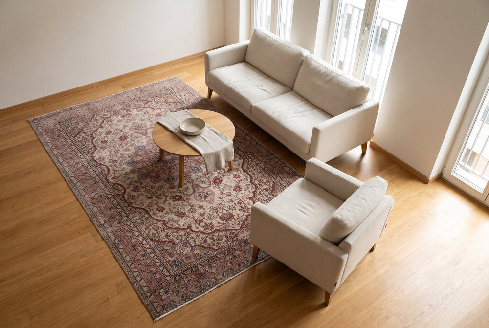 A cream and rose Isparta rug under a round oak coffee table with a linen sofa and armchair, seen from high above