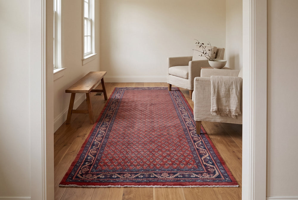 A long deep red vintage Arak rug with a small repeated motif and a navy border laid down a narrow sitting room beside a wooden bench and linen armchairs