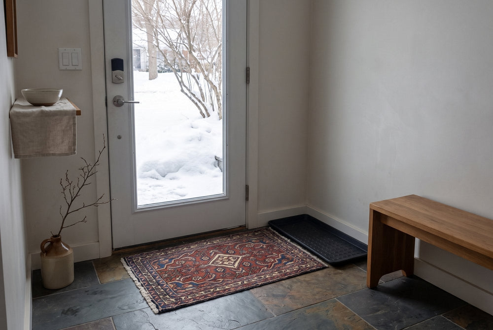 A small brick red vintage Hamedan rug with a diamond medallion lying inside a glazed front door with deep snow outside, beside an empty boot tray on slate tiles