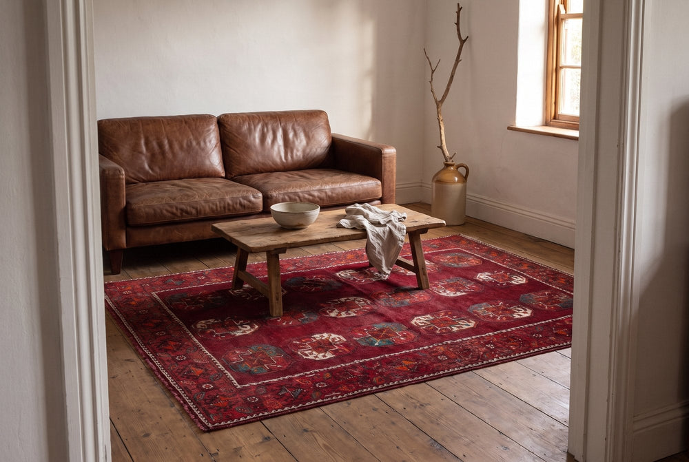 A deep red vintage Ghoochan rug with rows of octagonal guls under a wooden coffee table in a sitting room with a brown leather sofa, seen through a doorway