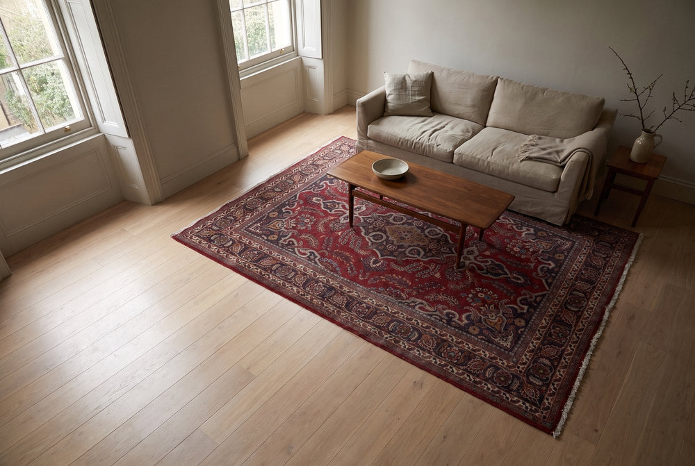 A red and navy Mashhad rug under a walnut coffee table in a bright living room with tall sash windows, seen from a high corner