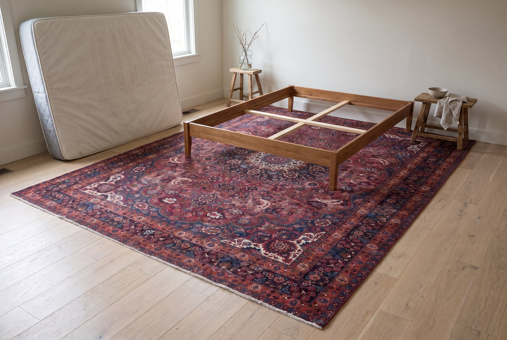 A burgundy vintage Dorokhsh rug with a rosette medallion and ivory corners lying flat under an empty wooden bed frame, the mattress leaning against the bedroom wall