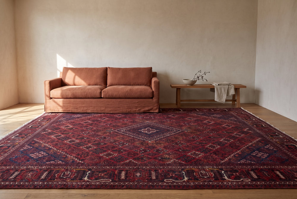 A burgundy vintage Joshaghan rug with a diagonal diamond lattice and a navy diamond medallion spread across a living room floor in front of a terracotta linen sofa and a wooden bench