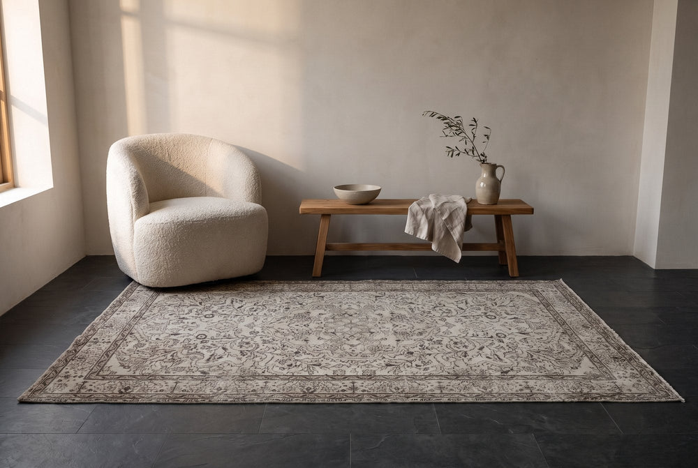 A pale gray and cream vintage Isparta rug with a faded charcoal medallion on a dark slate floor beside a boucle armchair and a wooden bench