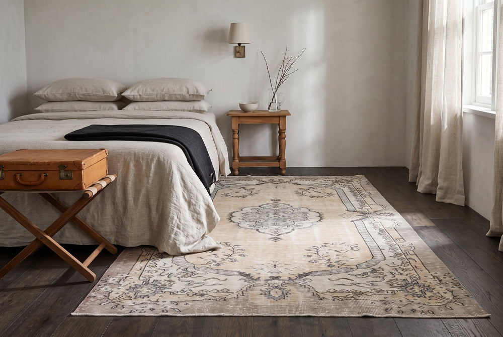 A pale cream vintage Isparta rug with a soft gray medallion laid across the foot of a guest bed on dark oak, with a leather suitcase on a folding luggage rack
