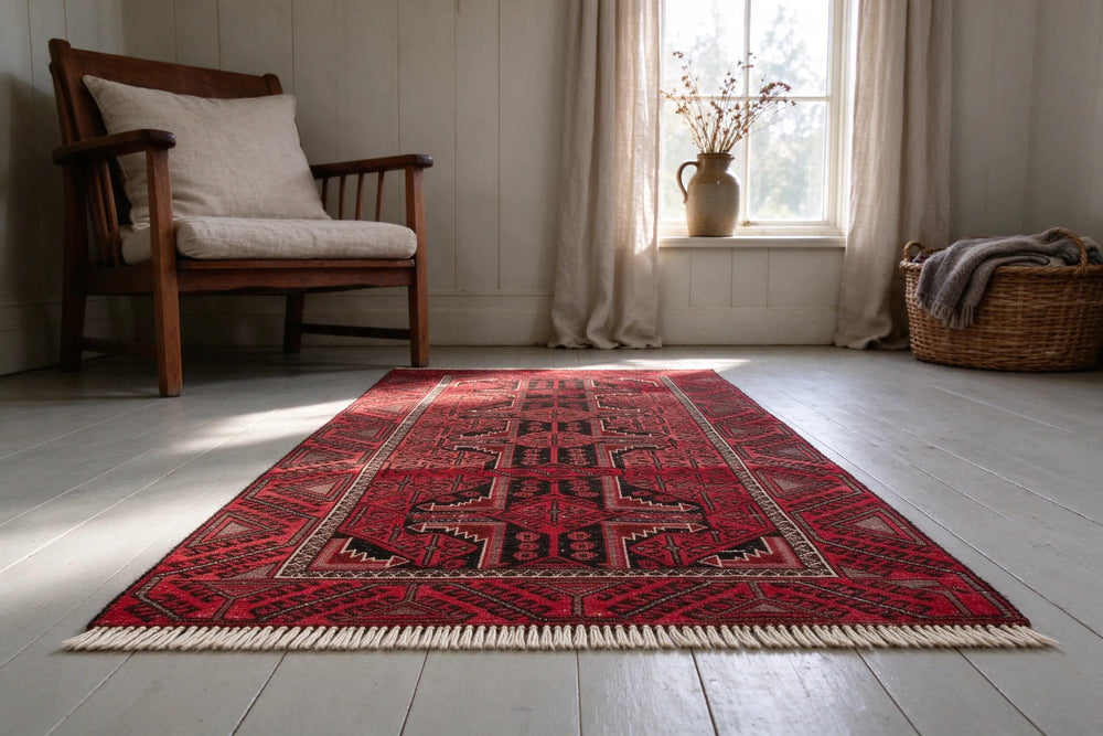 Low close view of the dense, short ivory fringe at the end of a deep red Baluch rug on pale gray painted floorboards, armchair behind