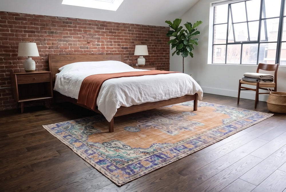 A queen bed with white linen on a faded terracotta and blue rug in a brick loft bedroom, the walnut nightstands off the rug.