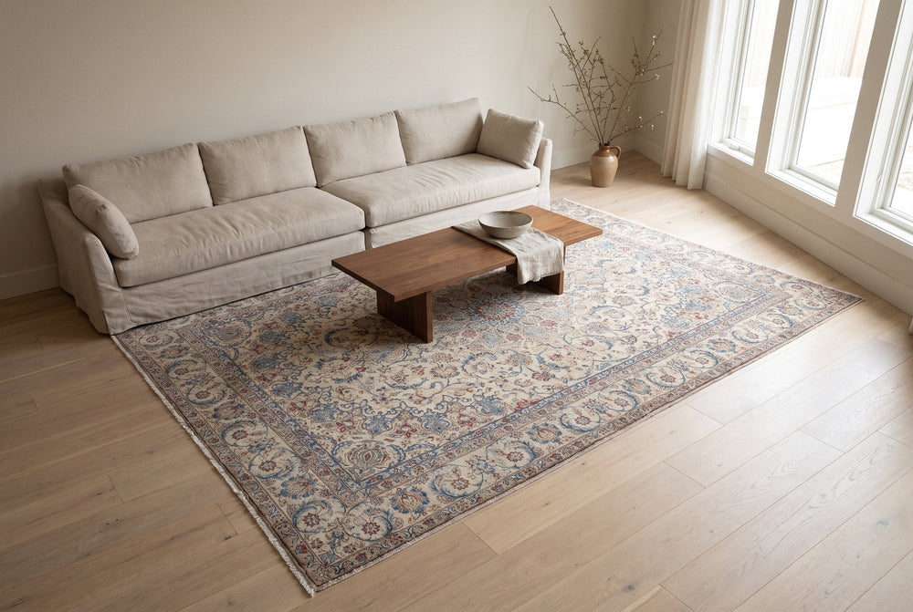 A cream vintage Nain rug with blue and red floral scrolls laid in a bright living room in front of a long linen sofa, with a walnut coffee table on it