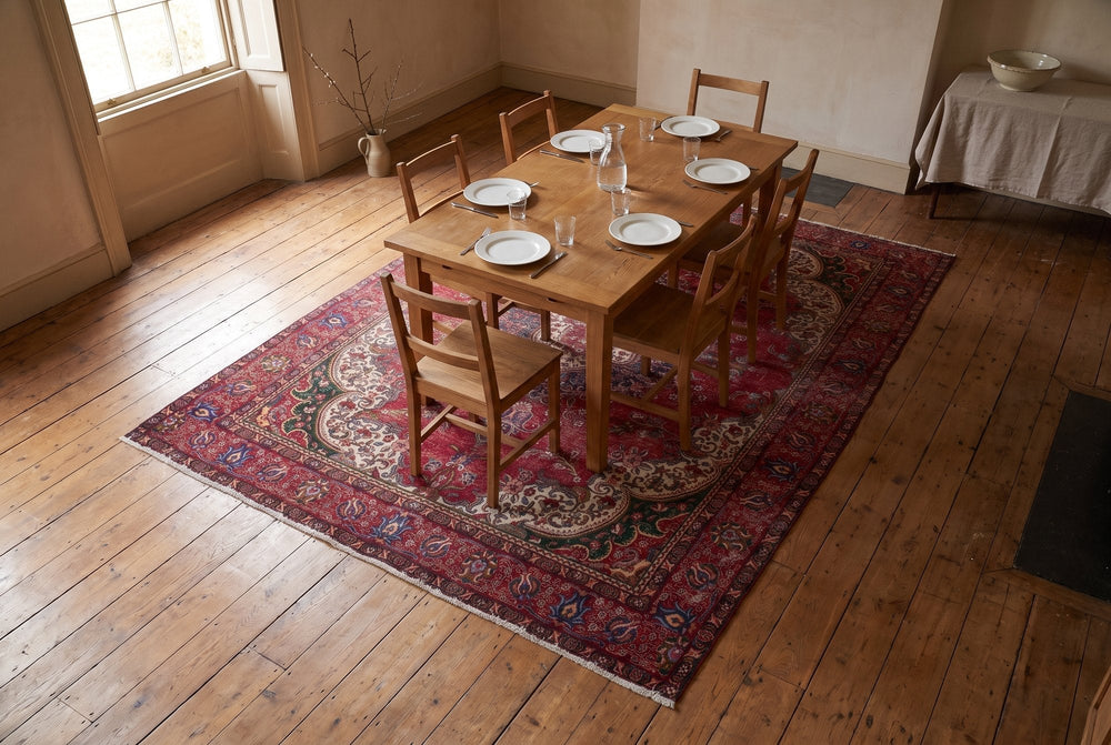 A red Tabriz rug with cream lobed corners under an oak dining table set for six, seen from above