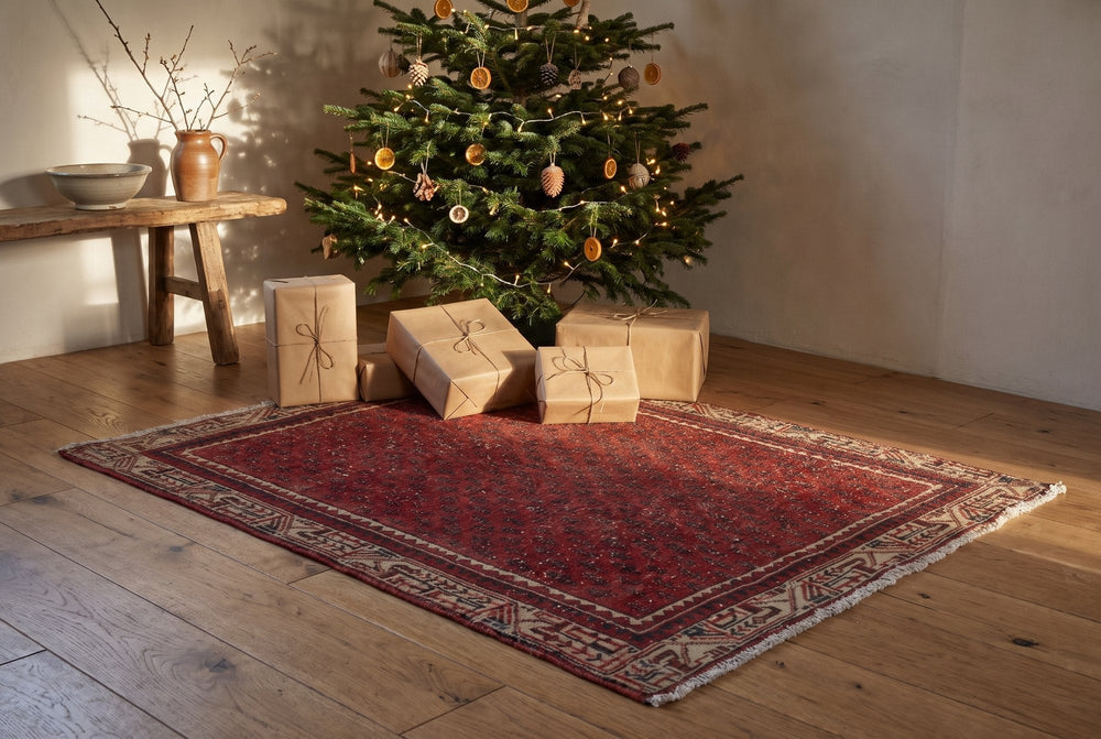 A small red vintage Arak rug with a tiny all-over repeat and a beige geometric border under a decorated fir tree, with presents in plain kraft paper