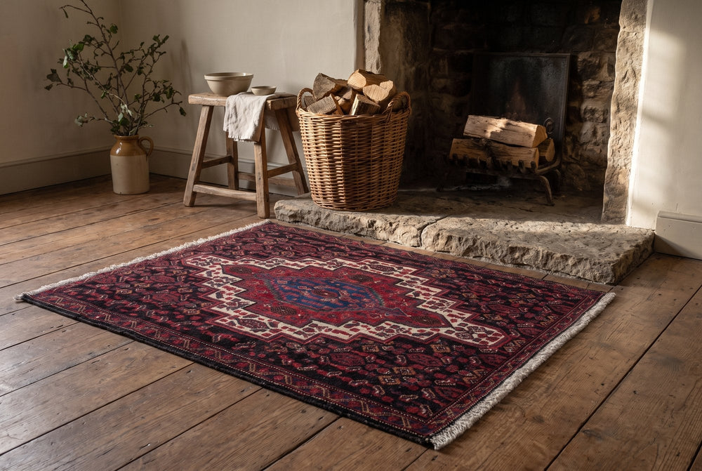 A small burgundy Sanandaj mat with an ivory-outlined medallion and dark blue center on pine boards in front of a stone hearth